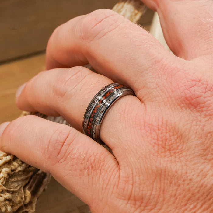 A close-up of a hand wearing The Makoshika, a wide silver Tungsten Carbide ring featuring dark and amber-colored inlays, resting on a textured, light-colored object.