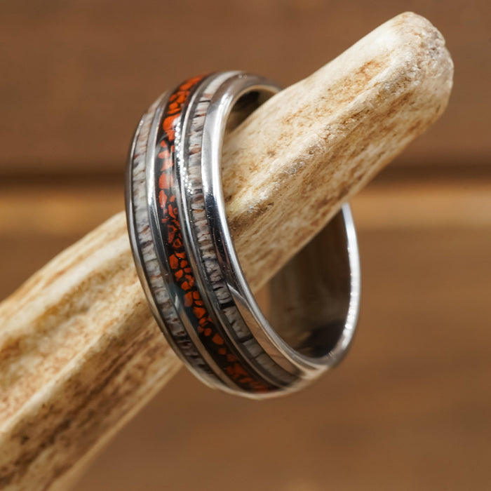 The Makoshika, a Tungsten Carbide ring featuring a wood-like inlay and striking orange accents, rests on a beige, textured antler against a softly blurred brown background.