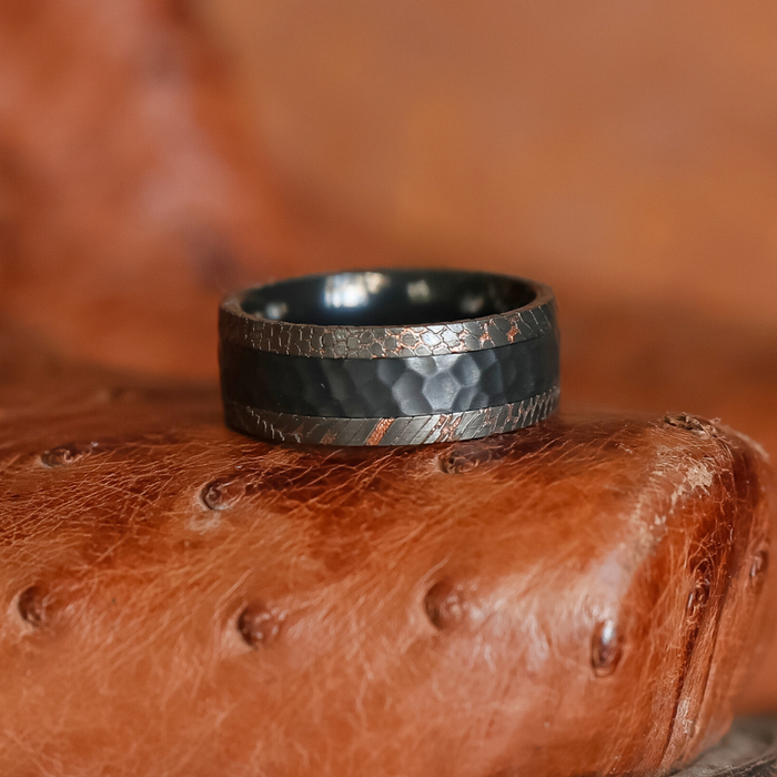 The Mamba, a black hammered metal ring featuring a textured band reminiscent of a rugged men's wedding band, rests on a brown leather surface with visible grain and dimples.