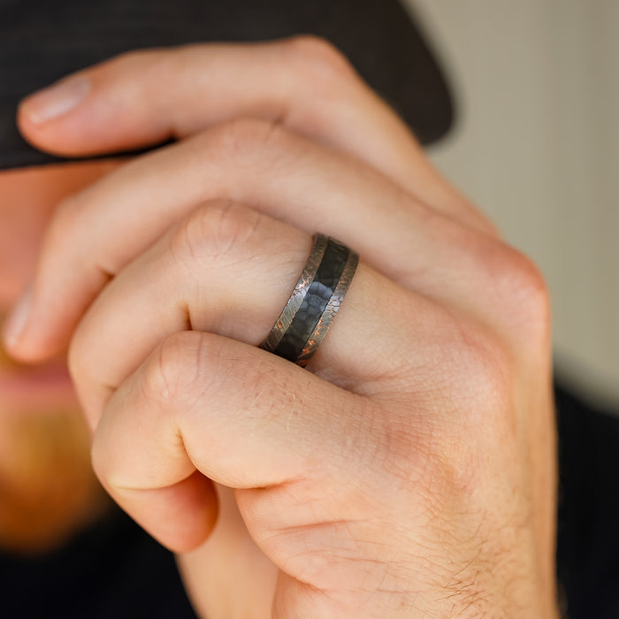 A close-up of a hand wearing The Mamba, a dark hammered-texture men's wedding band, on the ring finger. The person is partially covering their face with their hand and is wearing a black hat.