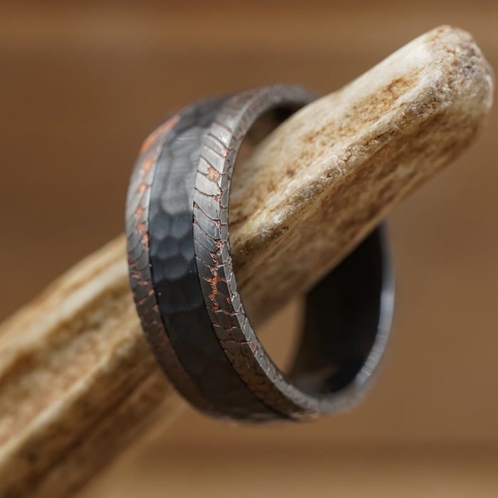 The Mamba, a dark, textured men’s wedding band with copper accents, rests on a light, rough antler against a blurred brown background.