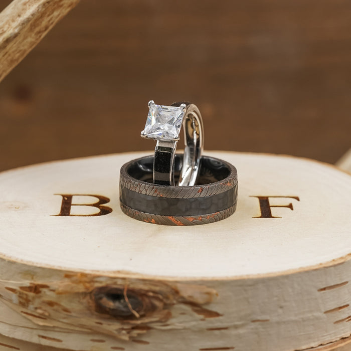 Two rings rest on a wooden surface engraved with B and F. One ring is silver with a square gemstone, while the other is The Mamba—a striking men's wedding band crafted from black zirconium and featuring a textured pattern.