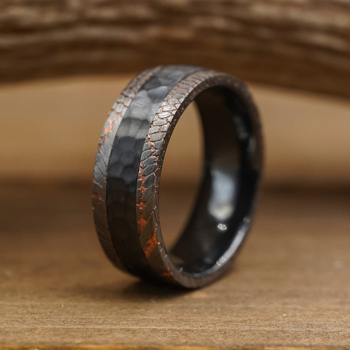 The Mamba, a dark hammered metal men's wedding band featuring a textured, reddish-brown pattern along its edges, sits upright on a wooden surface with a softly blurred wooden background.