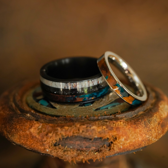 The Manchester & Patina Couples Set is elegantly displayed on a rustic surface against a blurred brown backdrop—one ring showcases striking black tungsten carbide with a silver and blue inlay, while its counterpart glows in silver accented by a unique brown and blue patina copper inlay.