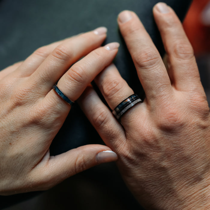 Two hands wearing simple rings gently touch against a dark background, symbolizing connection and partnership. The left hand wears The Manchester—a thinner blue band from The Manchester & Patina Couples Set—while the right hand showcases Patina, a thicker black tungsten carbide ring, beautifully highlighting their unique bond.
