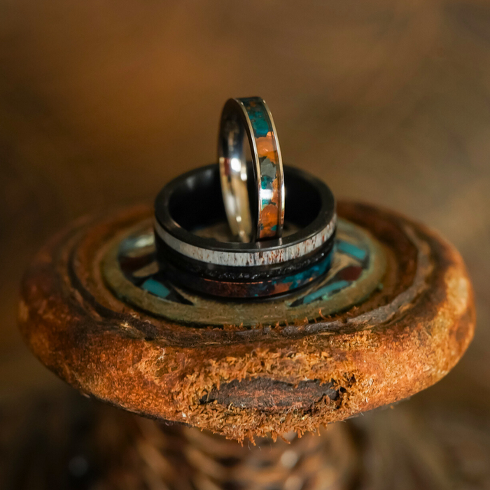 The Manchester & Patina Couples Set, featuring two rings with vibrant inlays—including a striking patina copper inlay—rests on a round, textured wooden surface accented with blue and brown tones. The background is softly blurred with warm brown hues.