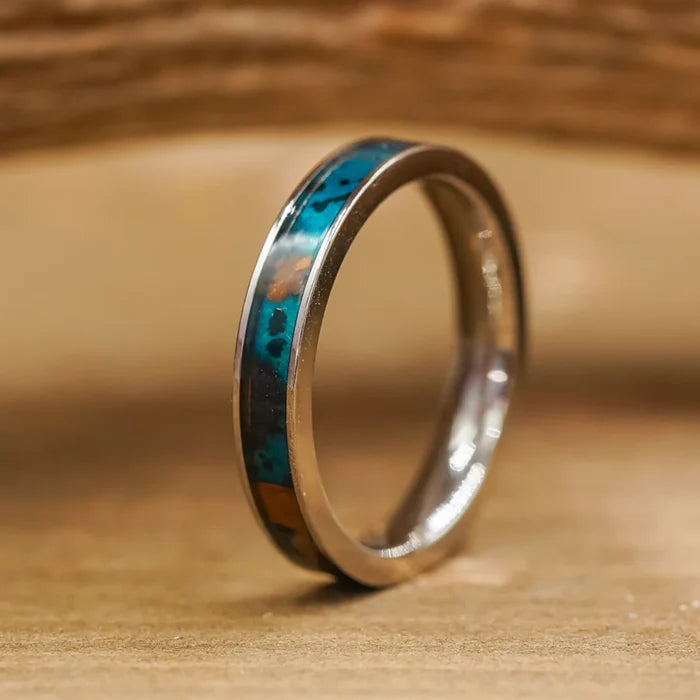 A stunning silver ring from The Manchester & Patina Couples Set, featuring a thin inlay of turquoise and brown stone, stands upright on a wooden surface with a softly blurred wooden background—its delicate design makes it an exquisite addition to any couples ring collection.