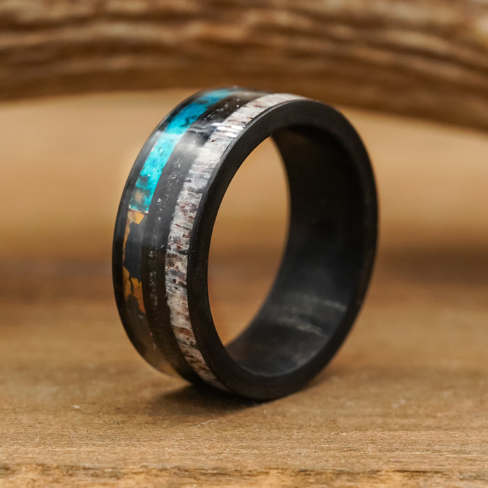 The Manchester is a black men's antler ring featuring two striking inlays: one of turquoise and another that resembles white and brown stone or wood, shown upright on a wooden surface with a softly blurred wooden background.