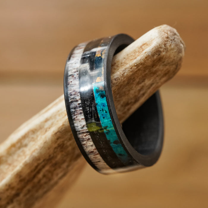 A close-up of The Manchester men’s antler ring, featuring black edges and vibrant inlays of turquoise, patina copper accents, and metallic bands, displayed on a textured light brown antler with a softly blurred brown background.