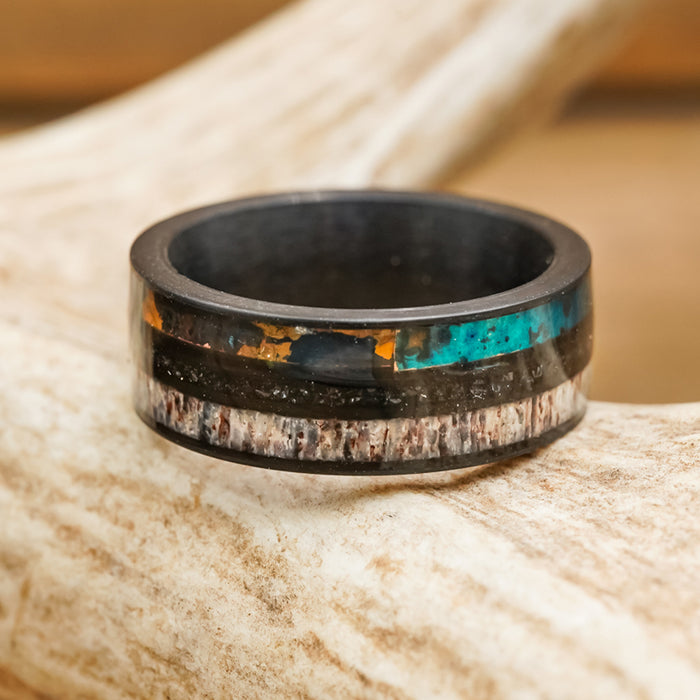 The Manchester is a men's antler ring with a black band featuring inlaid strips of turquoise, brown, and white materials, displayed on a light, textured surface reminiscent of antler.