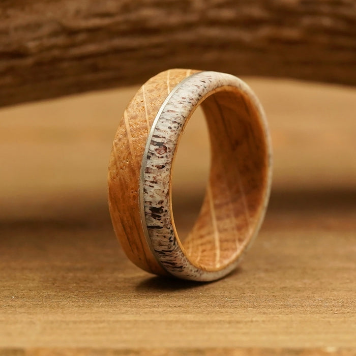 The Mount Amos, a wooden Whiskey Barrel Ring featuring a light brown color, dark speckles, and carved diagonal lines, stands upright on a wooden surface with a blurred wooden background.