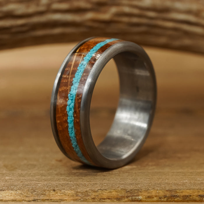 The Mount Vernon ring, crafted from silver tungsten carbide with a polished finish, showcases an inlay of brown wood and a thin stripe of crushed turquoise, elegantly displayed on a wooden surface.