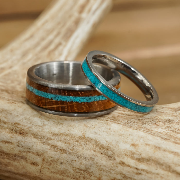 Two rings rest on a light antler. The larger ring, known as The Mount Vernon, features silver metal with inlays of wood and crushed turquoise, reminiscent of a whiskey barrel ring. The smaller ring is a sleek tungsten carbide band with a single turquoise inlay.