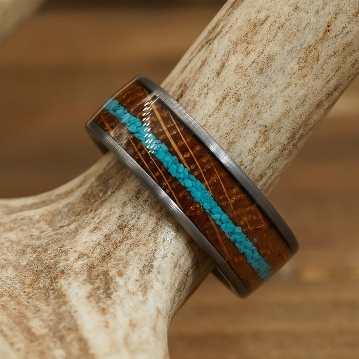 The Mount Vernon ring, featuring a brown wood-inspired tungsten carbide band with a vivid blue crushed turquoise inlay and polished metal edges, rests atop a light antler against a wooden backdrop.