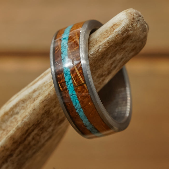 A close-up of The Mount Vernon ring, crafted from a whiskey barrel and inlaid with brown wood and crushed turquoise, displayed on textured antler with a softly blurred wooden background.