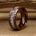 The Mount Vinson, a copper ring featuring a hammered texture and a striking white, wood-inspired inlay, stands upright on a wooden surface, reminiscent of a hammered antler ring. Its distinctive design makes it an ideal men's wedding band, set against the rustic backdrop of blurred wooden planks.