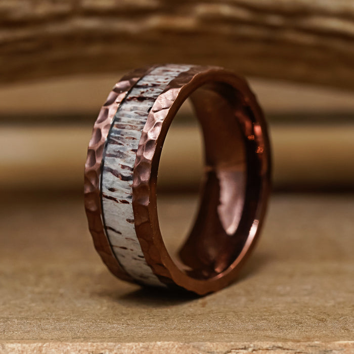 The Mount Vinson, a copper ring featuring a hammered texture and a striking white, wood-inspired inlay, stands upright on a wooden surface, reminiscent of a hammered antler ring. Its distinctive design makes it an ideal men's wedding band, set against the rustic backdrop of blurred wooden planks.