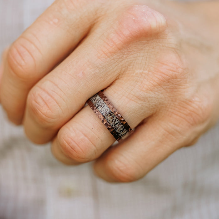 A close-up of a hand wearing The Mount Vinson, a dark, textured men's wedding band, on the middle finger. The background is softly blurred, revealing light, striped fabric.