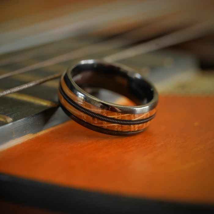 The Nashville, a dark, metallic Tungsten Carbide ring, rests atop the body of an acoustic guitar. Its sleek surface captures the cool allure of a guitar string ring, while the visible strings and wood grain in the background complete this harmonious composition.