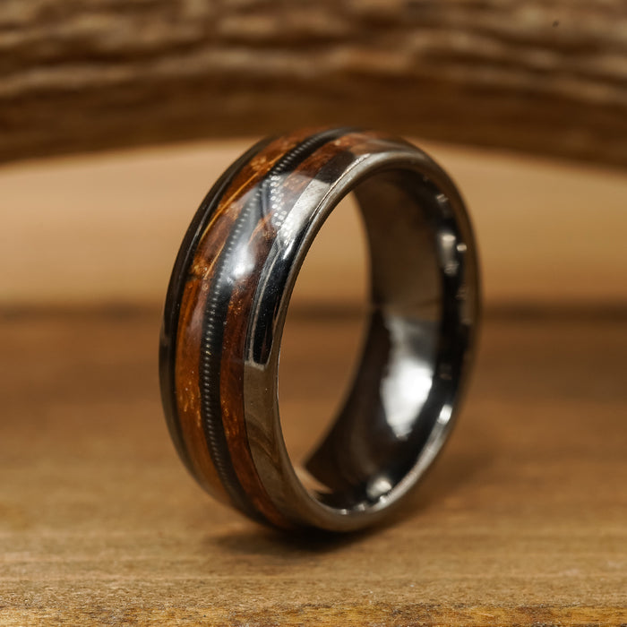 The Nashville, a Tungsten Carbide ring featuring a polished dark brown whiskey barrel inlay and a subtle grooved design, rests on a wooden surface with a softly blurred wooden background.