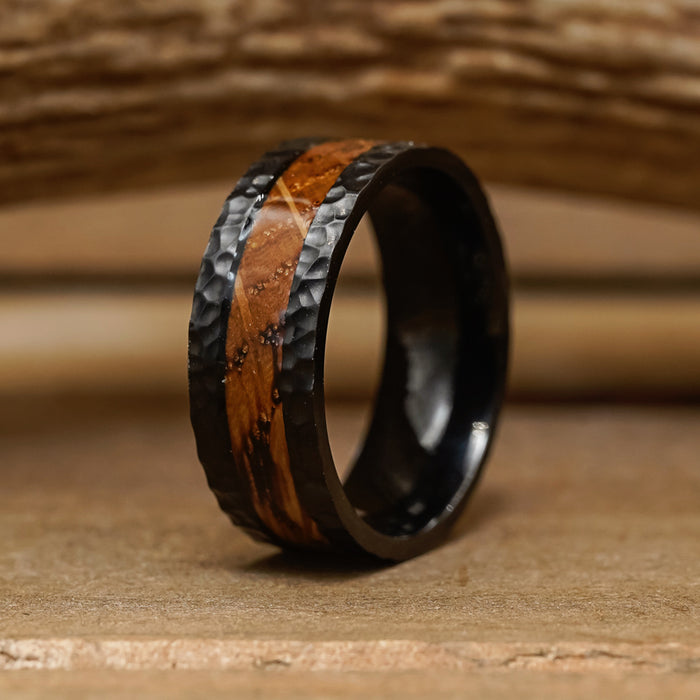 Introducing The Nickerson: a striking men's wedding band, this hand-hammered ring features a black titanium band with a unique hammered texture and a central inlay of polished brown wood grain, elegantly resting on a wooden surface.