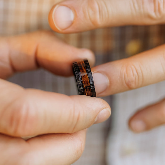 A person holds The Nickerson, a black titanium ring with a brown wood inlay, between their thumb and index finger, with a plaid shirt softly blurred in the background—an elegant men's wedding band that seamlessly blends modern style with natural elements.
