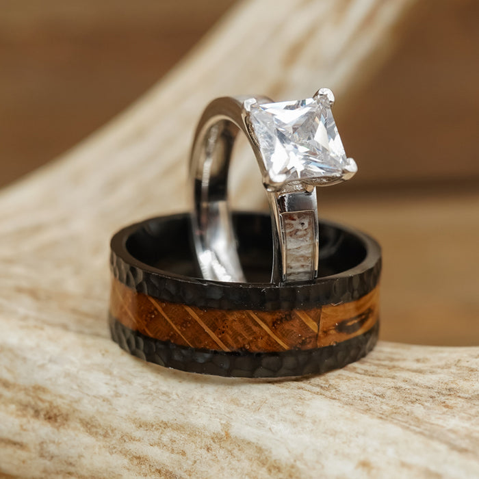 A silver ring featuring a square-cut clear gemstone is stacked above The Nickerson, a black and brown wood inlay titanium ring, both elegantly displayed on a light-colored antler.