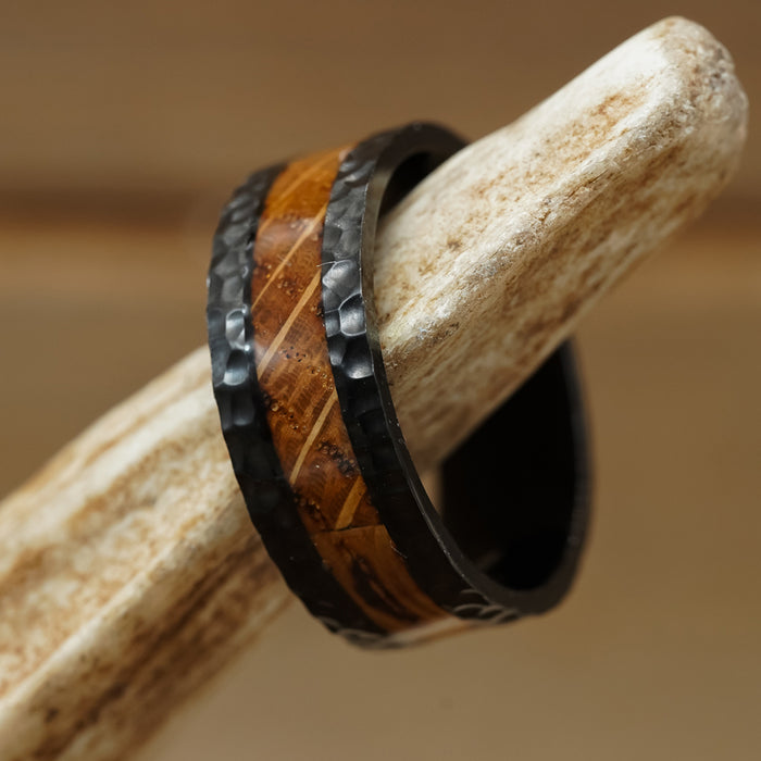 The Nickerson, a men's wedding band featuring a wood-like brown inlay and hand-hammered black metal edges, is displayed on a textured beige antler.