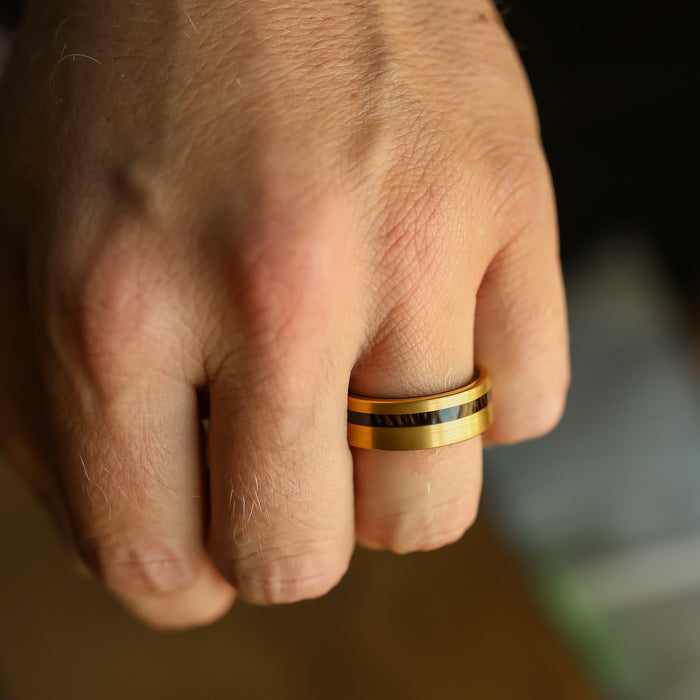 A close-up of a hand wearing The Oswald—a gold tungsten carbide ring featuring a dark center stripe reminiscent of a Wenge wood ring—highlighting skin texture and fine hair.