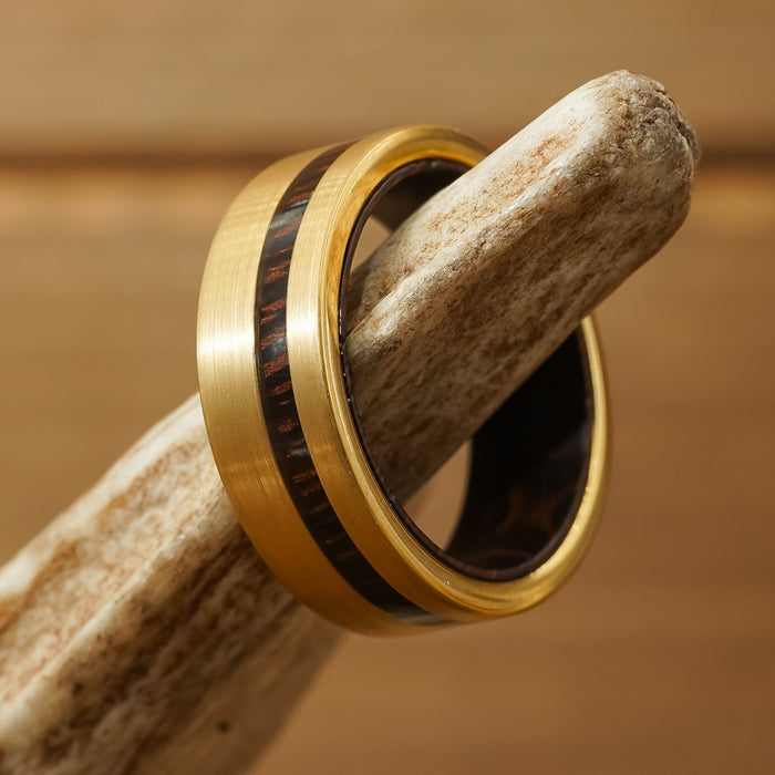The Oswald, a gold tungsten carbide ring featuring a dark Wenge wood inlay, rests on a light-colored, textured branch against a soft brown background, highlighting its elegance as a men's wedding band.