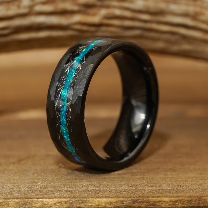 The Palo Verde, a black tungsten carbide ring featuring a faceted surface and a thin, luminous blue-green inlay, is displayed upright on a wooden surface with softly blurred wood grain in the background.