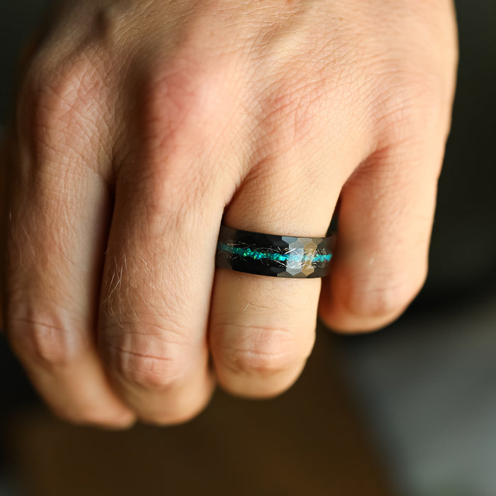 A hand wearing The Palo Verde ring—a black tungsten carbide band featuring blue-green marbled patterns—is shown in close-up, highlighting the intricate details and texture of the piece.