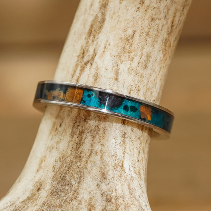 A women's copper stacking ring, The Patina, with a turquoise and amber-colored inlay, rests on a piece of antler against a blurred brown background.