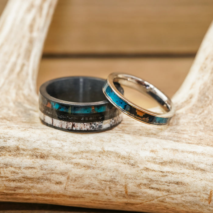 Two rings from The Patina collection, one a thicker patina copper band with a mix of blue and brown inlay, and the other a delicate women's stacking ring featuring a vivid turquoise stripe, rest on a light-colored antler. The background is blurred wood.