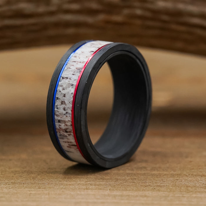 The Patriot ring, crafted from black carbon fiber with a textured white center band and bordered by thin blue and red lines, rests upright on a wooden surface.