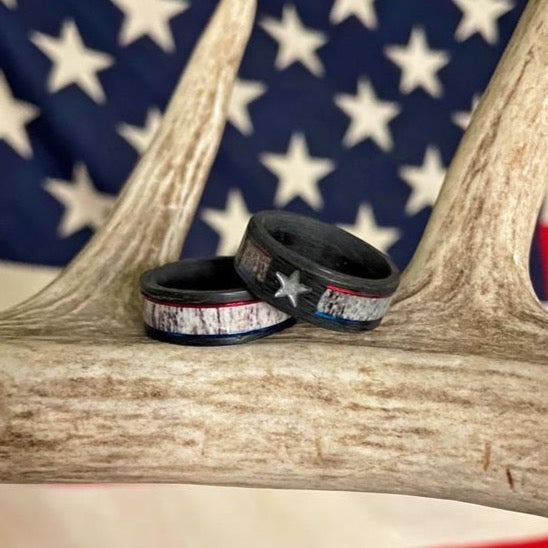 Two black carbon fiber rings from The Patriot collection, featuring American flag designs and a star, rest on an antler. A blurred U.S. flag in the background highlights these bold men's wedding bands—perfect for those who appreciate rugged style and patriotic details.