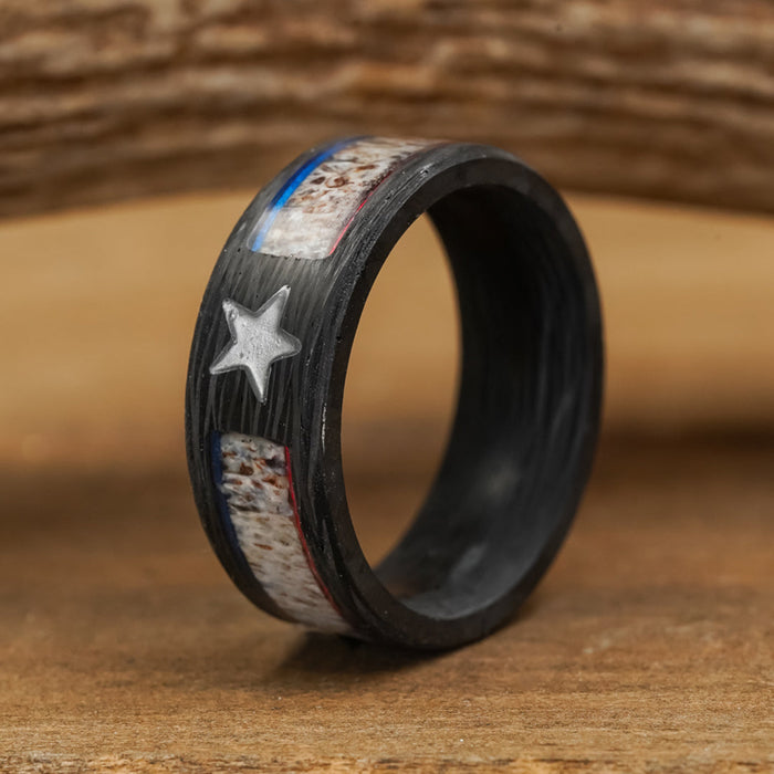 The Patriotic Star: A black carbon fiber ring featuring a silver star and red, white, and blue bands on its surface, displayed against a wooden background—the perfect men’s wedding band for those seeking a unique style.