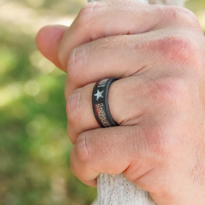 A close-up of a hand gripping a white stick, wearing The Patriotic Star men's wedding band—a dark ring featuring a white star with red, white, and blue accents—set against a blurred green background.