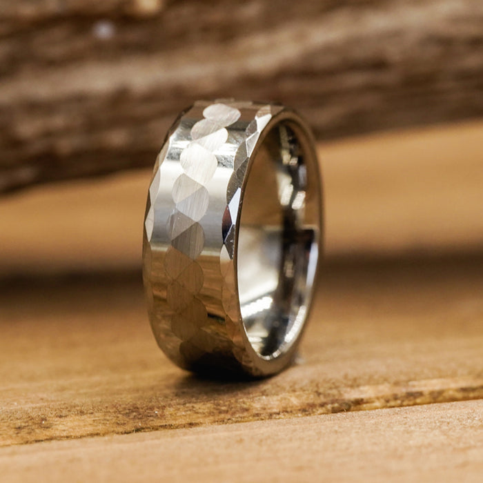 The Payson, a hammered silver ring featuring a faceted, textured 8mm band, sits upright on a wooden surface, catching the light against a softly blurred wood background.