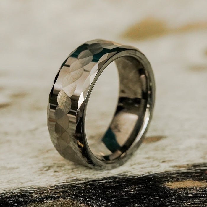 A close-up of The Payson, a metallic hammered silver ring featuring a faceted geometric pattern, resting upright on a textured surface of light and dark tones.