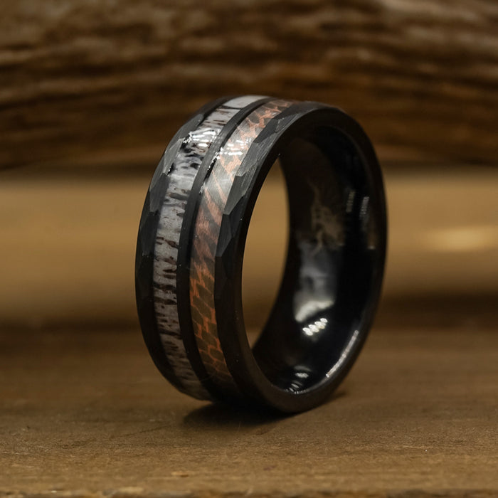 The Python, a black tungsten carbide ring with a textured angular design and an inlay that blends silver and copper tones, sits on a wooden surface with a blurred background.