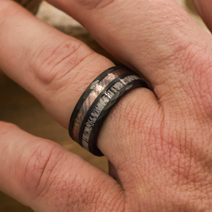 A close-up of a hand wearing The Python, a black tungsten carbide ring with metallic and textured inlays, shown on the ring finger.