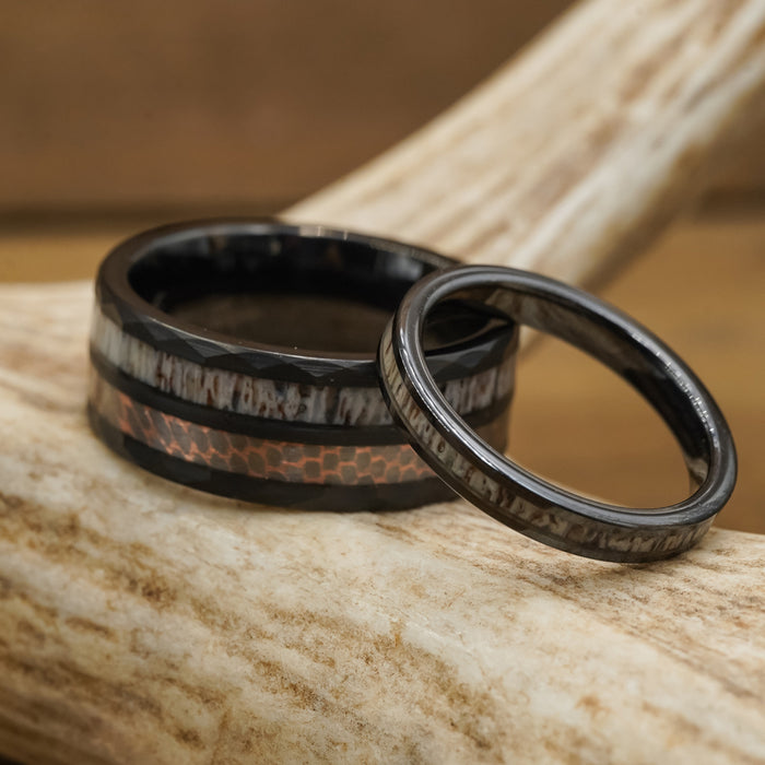 Two rings rest on a light-colored antler. The thicker band is The Python, a striking tungsten carbide ring with a black exterior and copper inlay, while the thinner ring features a dark, wood-like pattern that perfectly complements the natural elegance of the antler ring display.
