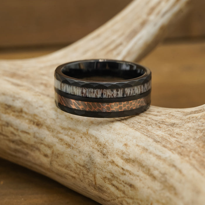 The Python, a black tungsten carbide ring featuring a patterned copper inlay and a light, textured band, rests on a piece of antler against a blurred brown background.