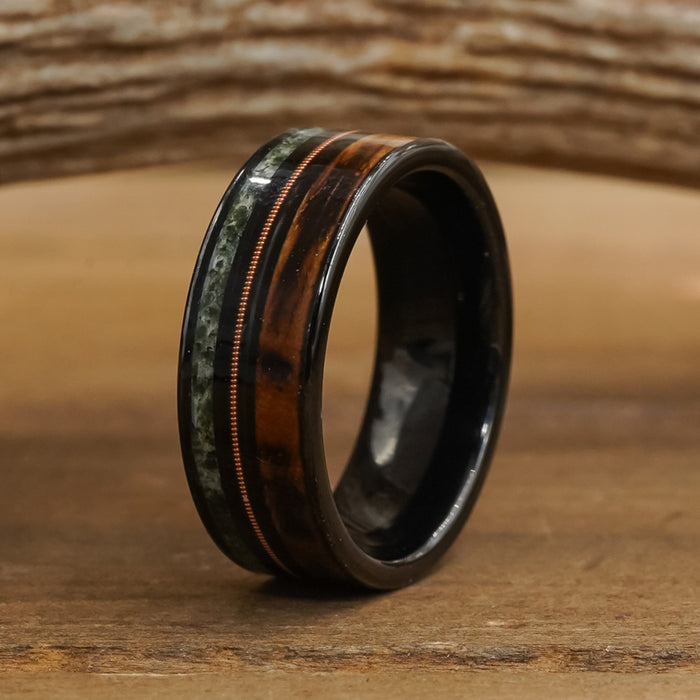 Introducing The Ricketts Glen: a black Tungsten Carbide ring featuring a polished brown wood inlay, a slim copper stripe, and a green moss agate accent, shown standing upright on a wooden surface with a rustic wood backdrop.