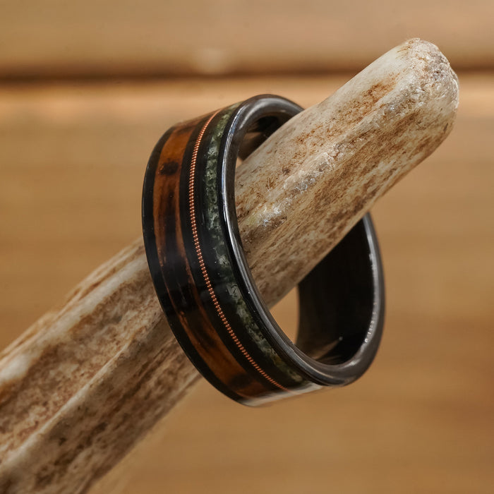 The Ricketts Glen, a black Tungsten Carbide ring featuring brown and green inlays with a thin copper stripe, rests on a textured, light-colored antler against a wooden background.