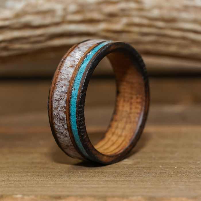 The San Gabriel, featuring a wide, light-colored textured band crafted from whiskey barrel wood and accented with a turquoise blue inlay, stands upright on a wooden surface against a softly blurred wood background.