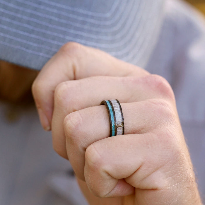 A person wearing a gray cap holds their fist near their face, showcasing The San Gabriel ring—crafted from whiskey barrel wood with a striking metallic and turquoise inlay—on their finger.
