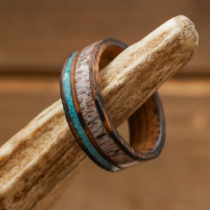 The San Gabriel, a wooden ring showcasing contrasting light and dark bands and accented with a vibrant turquoise inlay, is displayed on a piece of antler against a softly blurred brown background.