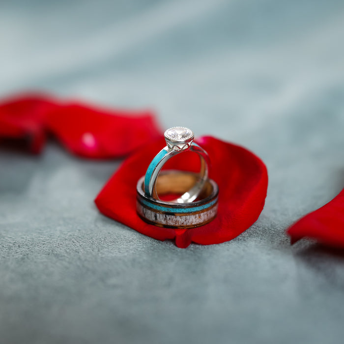 The San Gabriel, a ring featuring a delicate diamond, rests alongside a plain whiskey barrel ring on a red rose petal atop a soft gray surface.